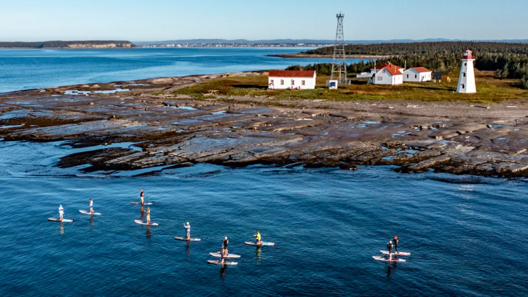 Groupe de personnes en paddle sur l'eau près d'un phare et de maisons sur la Côte-Nord, illustrant les activités récréatives en plein air dans la région.