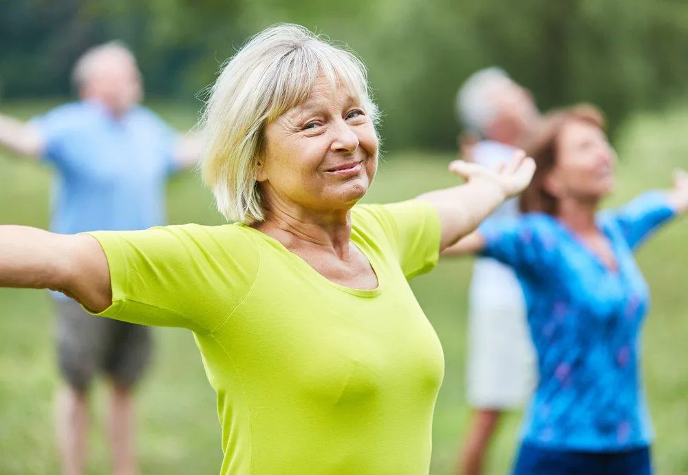 Femme d'âge mûr souriante en t-shirt vert, les bras ouverts, participant à une activité de bien-être en plein air avec d'autres personnes en arrière-plan, illustrant les services de santé mentale et de suivi à la Clinique Omicron.
