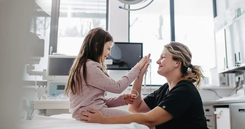Femme m&eacute;decin souriante interagissant avec une jeune patiente dans un environnement m&eacute;dical, illustrant l'approche humaine et centr&eacute;e sur le patient de la Clinique Omicron.