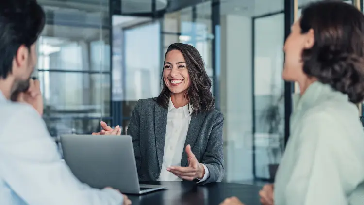 Femme souriante en réunion d'affaires, discutant avec des collègues autour d'un ordinateur portable, illustrant les services de santé en entreprise de Clinique Omicron.