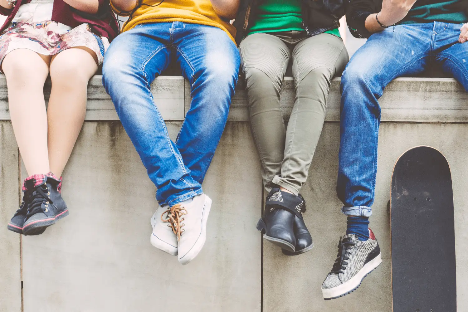 Group of friends sitting on a concrete ledge, showing legs in sneakers; a skateboard rests beside them.