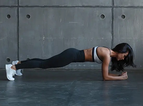 Woman performing a forearm plank in a gym, body straight with elbows on the floor against a concrete wall.