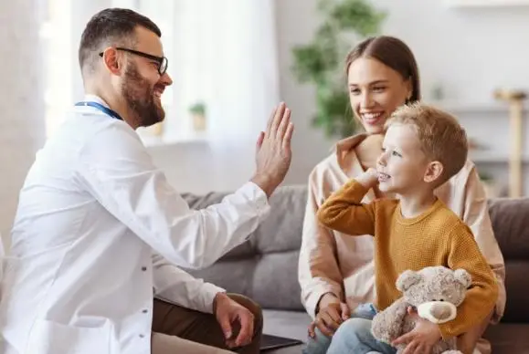 Pediatrician high-fiving a smiling child as the child's mother watches in a bright clinic lounge.